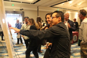 A Latino man in glasses and a black shirt points to a poster while talking to a smiling Italian woman