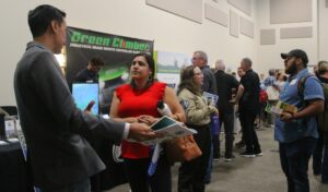 A Latino man in a suit and a Latina woman in a red shirt talk in the foreground while a crowd of various people mill about behind them, looking at posters and presentations