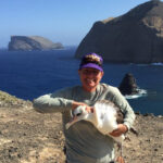 A white woman in a baseball cap smiles while holding a large seabird in her arms
