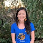 A young woman in a blue shirt poses smiling in front of a eucalyptus tree
