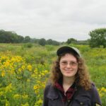 A young woman in a hat stands before a field of yellow flowers