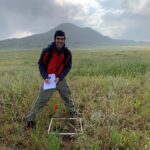 A smiling young man in a red jacket pauses while taking notes on a square patch of plants