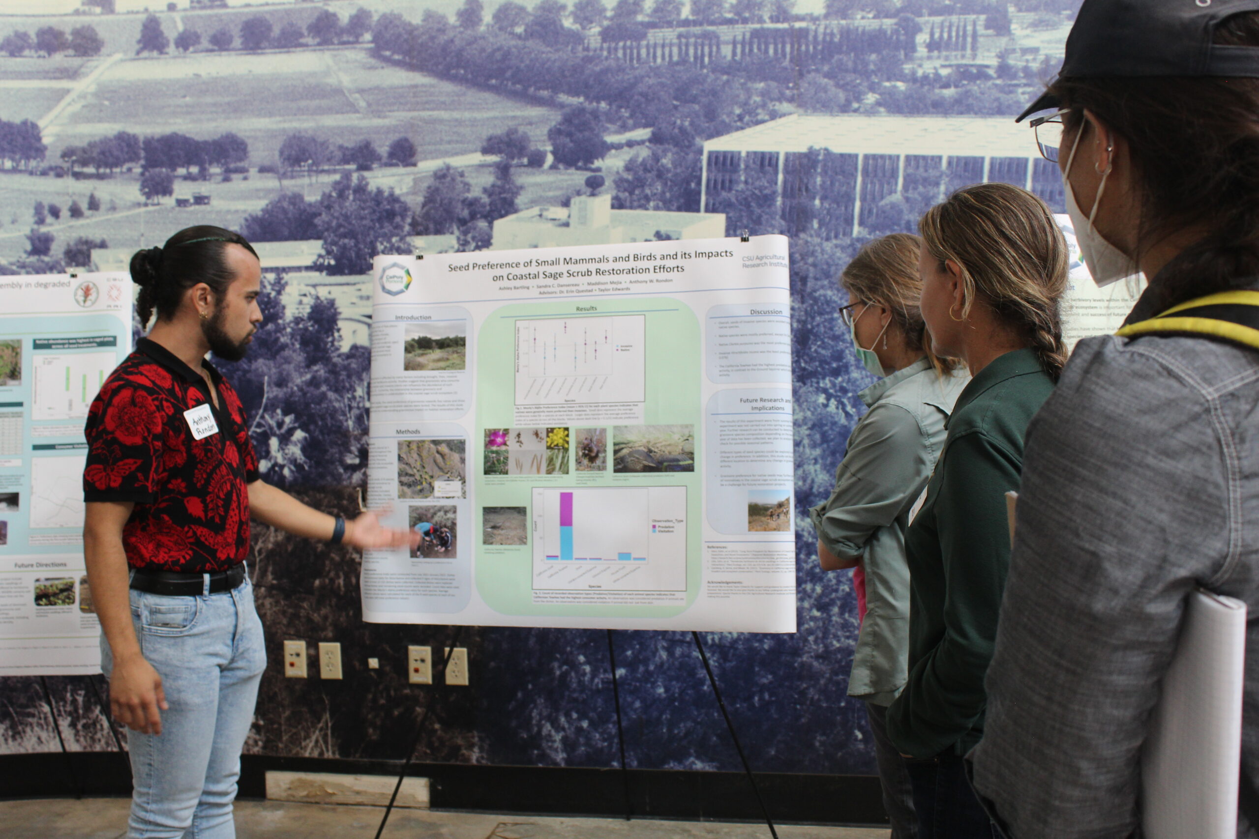 A young man with long dark hair swept into a top bun and beard in a black and red shirt stands to the left of a poster with dense text and pictures. He gestured towards it while three women in masks and dark clothes look attentively at him.