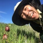 A woman in a sun hat leans close to a purple flower growing in a field with a huge open mouth smile