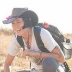 A woman with short black hair wears a grey brimmed hat white shirt and black pack. She leans over and laughs