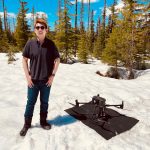 A man in a black polo shirt, dark glasses and jeans stands smiling with thumbs in jean pockets on a snowy field. At his feet is a black drone on a black tarp. Behind him are green pine trees under a blue sky. 