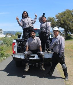 Four young people in work uniforms, one in a hard hat, stand or sit on the bed of a white truck filled with black garbage bags. They smile and make raise "peace sign" hands.