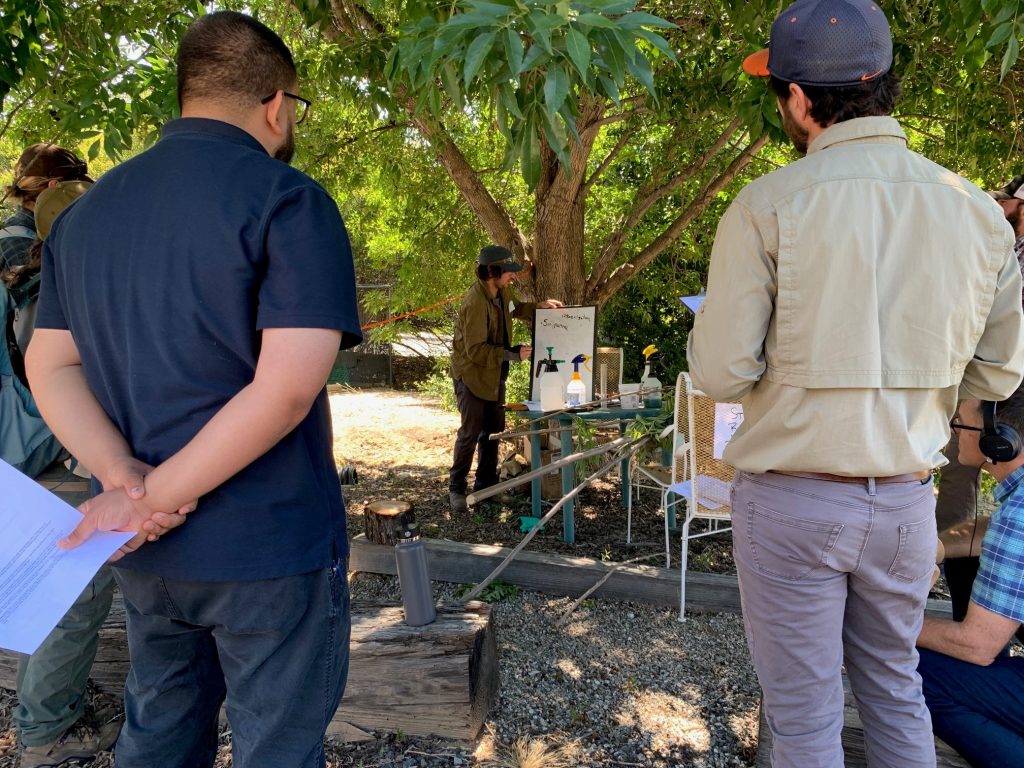 Two people face away from the camera and watch a man in a hat with charts showing herbicide calibration. 