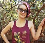 A woman with a purple undercut bob and black glasses smiles. She has one hand on a small guava tree and one hand on her hip. She wears a maroon tank top with the state of California on it. 