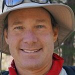 A man in a red kerchief and tan sun hat smiles into the camera