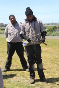 Two young men in work clothes and boots clean their tools in a grassy lawn next to an estuary