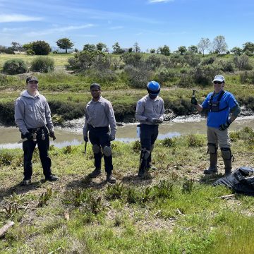 Four young men in hard hats and work clothes hold tools and stand in mud and grass by an estuary