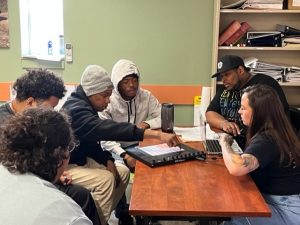 A group of young men and women site around a table and work together to write a song, writing ideas on paper