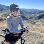 Smiling woman in sunglasses and a bike helmet sits on a bike on a sunny trail with rolling green and blue hills behind her