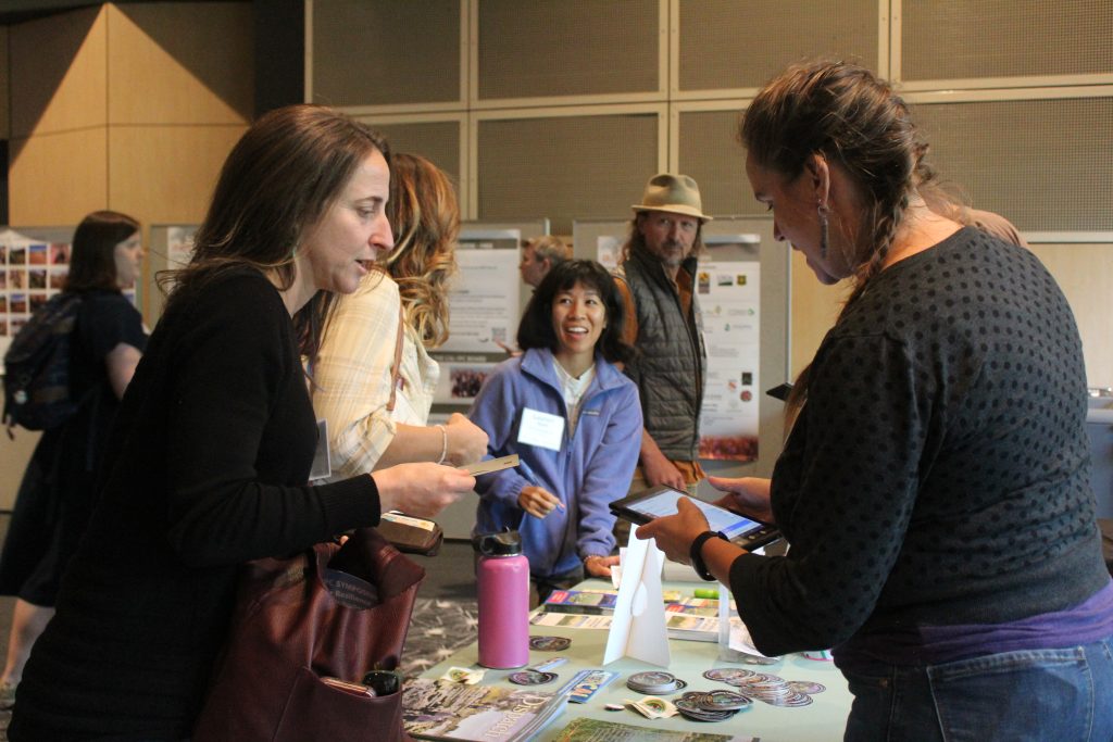 A woman at a table covered in magazines and brochures answers anther woman's question in a busy conference hall