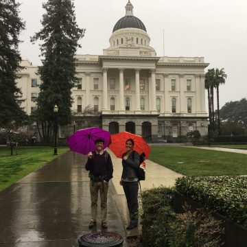 A smiling man and woman with purple and red umbrellas stand on wet sidewalk in front of the state capitol on a grey rainy day