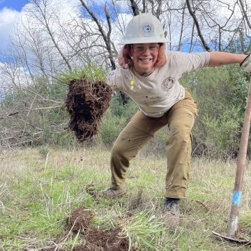 A grinning young woman in a white hard hat holds a fistful of harding grass.